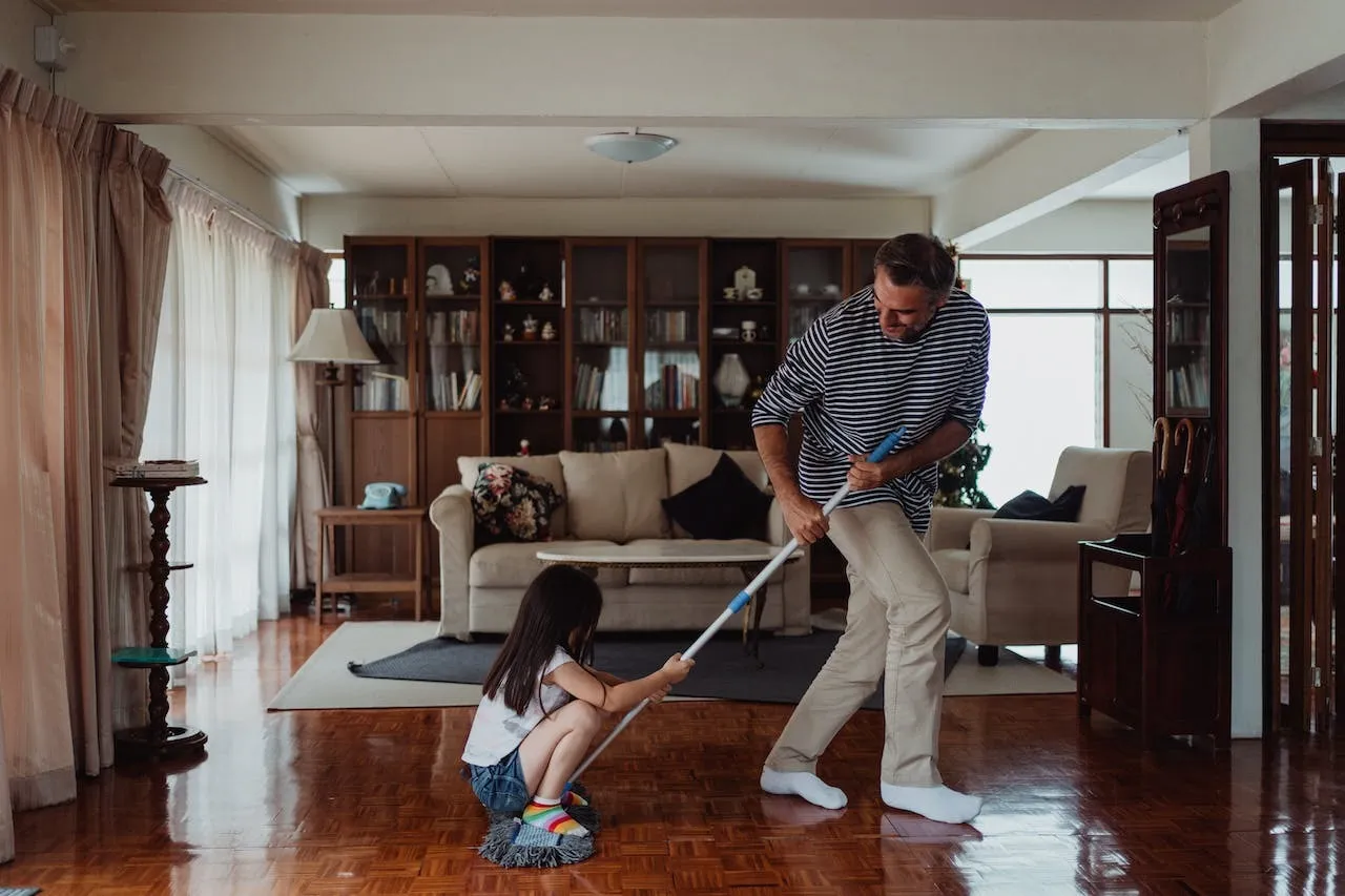 A man cleaning a hardwood floor with a little girl crouched and holding on to a mop
